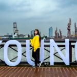 A young woman poses next to large letters spelling 'YOUNG' with an industrial waterfront backdrop.